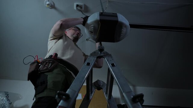 ServiceMan repairing garage door opener. Technician checks wires on ceiling mounted motor in residential garage.