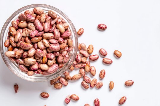 Pinto beans in a glass bowl, dry raw legumes spilled on a white background. Top view of healthy protein-rich food ingredient.