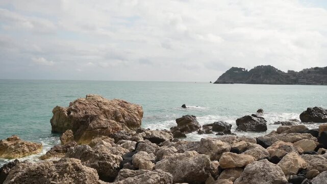 Waves gently crash against a rugged shore filled with various sized rocks and boulders, leading to a vast expanse of calm sea