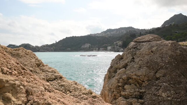 View of a rugged coastline with large rocks in the foreground and a town nestled on a hillside overlooking the sparkling blue sea