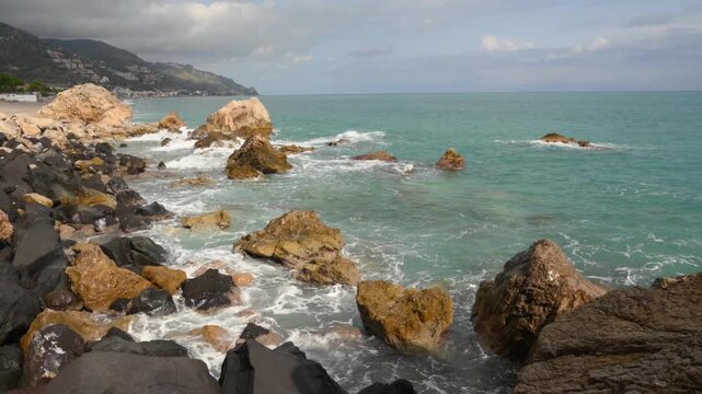 Turquoise ocean waves gently lap against a rugged coastline dotted with large boulders and smaller rocks, with a distant town nestled on a hillside