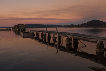 Obraz premium Wooden pier leading to a calm lake at sunset with warm orange sky, peaceful water reflections and distant hills, tranquil nature landscape perfect for travel, relaxation, serenity and scenic backgroun
