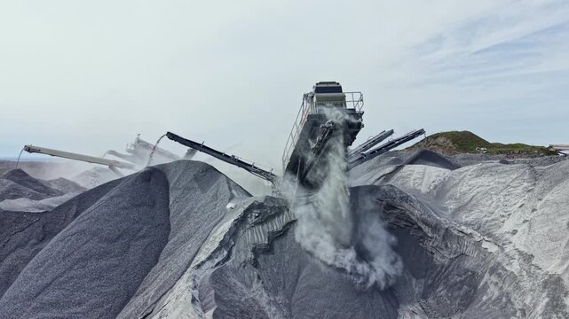 Heavy machinery works in a granite quarry, moving and processing stone. Large piles of crushed granite are formed as equipment moves around the site. Dust fills the air during operations.