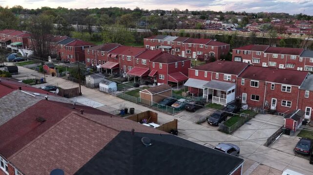 Aerial flyover of Baltimore suburb in spring, with classic red brick row houses, blooming pink trees, quiet residential streets and dense neighborhood layout with distant highway traffic. Wide shot.