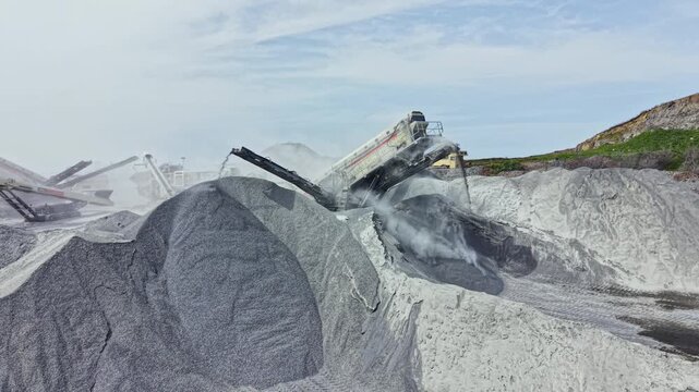 Machines work in a granite quarry to crush and process stone. Dust rises as large piles of granite form. Workers manage operations at the site during daytime.