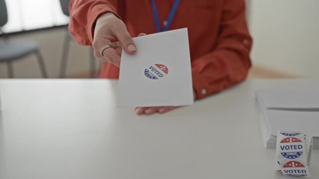 Woman young hispanic handing ballot envelope across a table with visible hand and roll of voted stickers at building desk; civic duty participation pride.