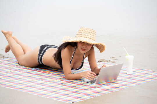 Southeast Asian woman relaxes on a sandy beach, enjoying a vibrant cocktail and a checkered blanket, wearing a black bikini and sunglasses, capturing a serene coastal moment.