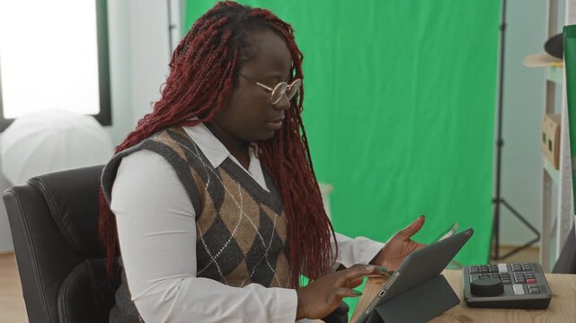 Woman with long red locs and glasses taps a tablet screen with index finger while seated at desk beside control panel and studio lights in a studio with green screen; focus productivity creativity.