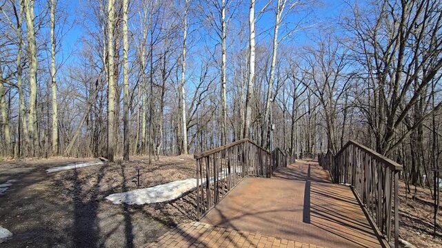 A wooden deck with wooden railings leads to a bare winter forest on a clear sunny day, when the snow has not yet fallen