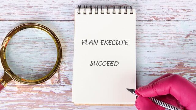A hand wearing a red glove writes the words Plan Execute Succeed on a white spiral notebook with a pen