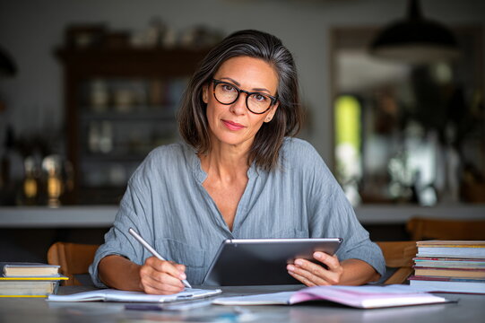 Mature Hispanic woman with glasses studying intently at a table, focused and engaged with tablet