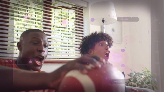 Two men sitting on couch, watching off-frame sports play, repositioning football, cheering