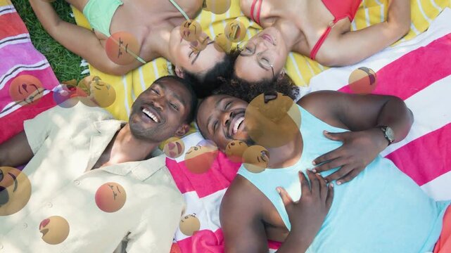 Four friends lying on grass towels, overhead cam sparking emoji overlays, laughing for marketing