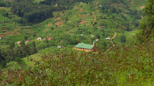 Rural church positioned within terraced tea plantation landscape in Bwindi highlands with scattered houses and winding paths across rolling hills in a cultivated agricultural environment
