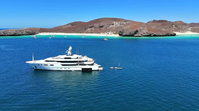 4K drone orbiting a yacht in clear calm water at Playa Balandra in La Paz, with the beach and coastline in the background.