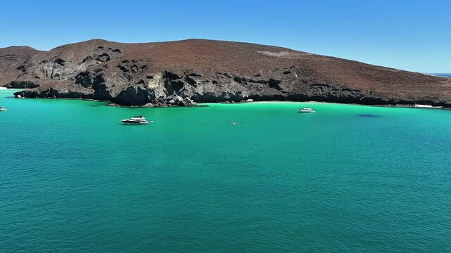 4K drone following birds over clear water at Playa Balandra in La Paz, with rocky cliffs, beach, and yachts in the background.