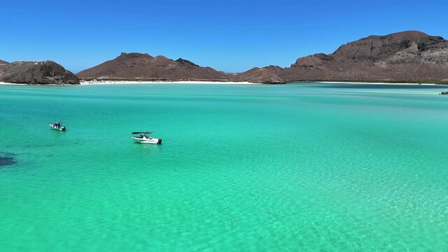 4K drone flying low over clear shallow water at Playa Balandra in La Paz, passing boats and moving toward the beach and mountains.