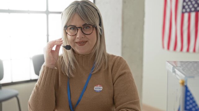 Woman wearing headset touches microphone and shows i voted sticker at a voting desk in building; civic duty friendly.