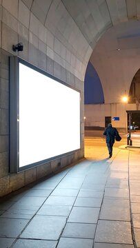 Blank White Horizontal Lightbox Billboard Mockup On Stone Wall Under Arched Urban Walkway At Night With Pedestrian Walking Away On Paved Sidewalk