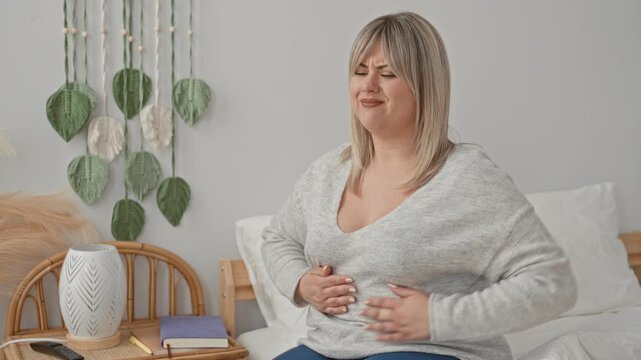 Woman pressing abdomen with hand, wincing and seated on bed in a bedroom with bedside lamp, rattan chair, journal and macrame wall hanging; discomfort nausea.