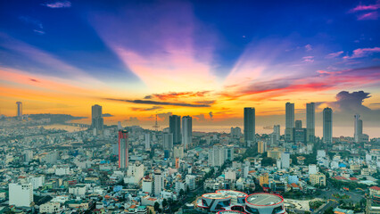 Obraz premium A serene sunrise view of Nha Trang city, Vietnam, where the soft orange sun rises above the coastal skyline. Layers of high-rise buildings and residential neighborhoods stretch toward