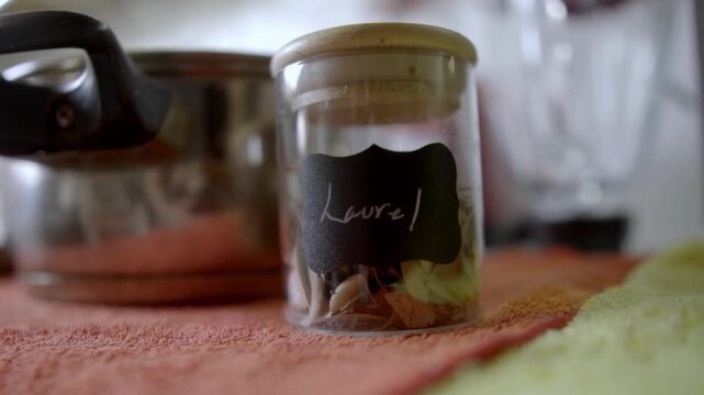 Closeup of glass jar containing garlic cloves placed on kitchen table with utensils in background showing food ingredient storage and cooking setup