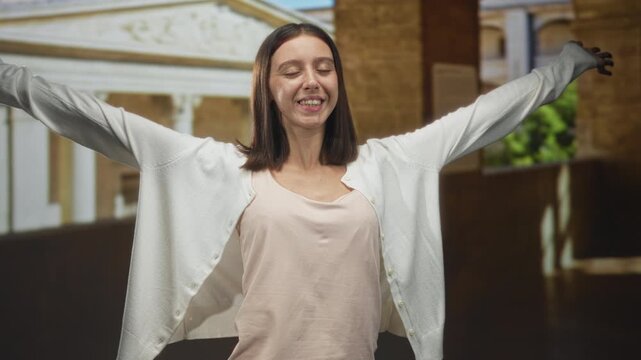 Young woman in white cardigan with arms outstretched and eyes closed, taking a deep breath inside a building; serenity freedom breathing.