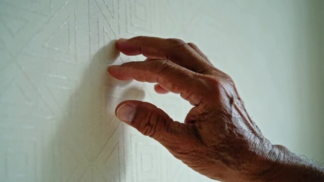 Close-up of an elderly Black man&rsquo;s hand tracing embossed geometric wallpaper. Soft light highlights texture, touch, and sensory interaction in an elegant interior.