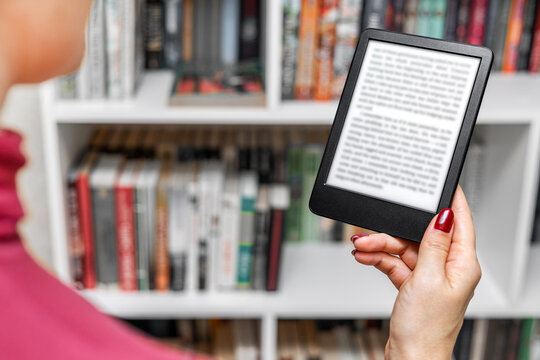 a woman holds an e-book in her hand. A woman reads an e-reader near a bookcase. an e-book against the background of paper books. A woman reading an e-book at home. 