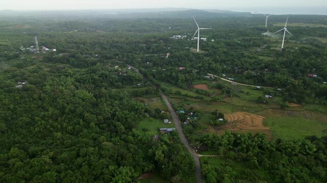 Aerial flyover of a winding road traversing lush farmlands with wind structures in the distance. Ideal for projects on sustainable energy or rural landscapes.