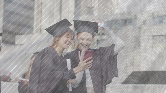 Two graduates leaning toward phone showing message on campus plaza, fixing caps, for graduation