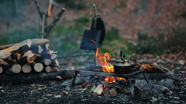 Campfire burning with a tea kettle placed next to a metal grill and stacked firewood in the background during a daytime outdoor camping session.