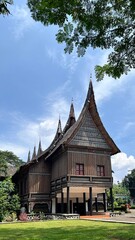Obraz premium Traditional Indonesian house with curved, pointed rooflines rises against a bright blue sky, features intricate decorative patterns on its wooden exterior and stands elevated on pillars.