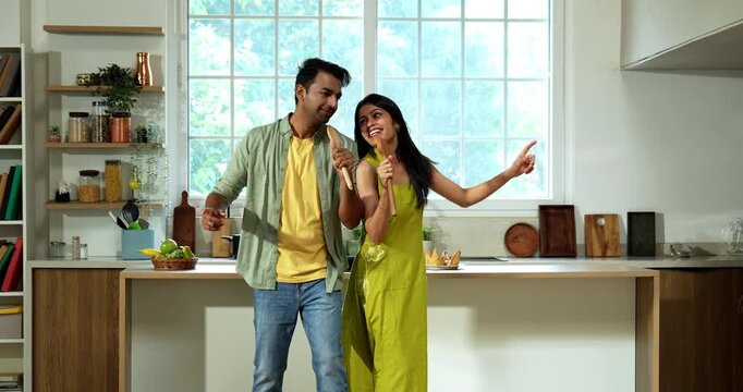 Indian asian young married couple singing song standing back to back with spatula as mic and dancing in modern kitchen having fun together during weekend morning at home in natural light