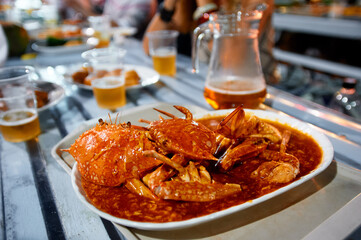 Steaming chili crab served in a rich red sauce on a communal table, surrounded by plastic cups of...