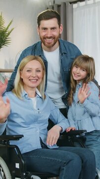 Home interier family portrait of charming joyful adult handicapped mother in wheelchair ,likable positive bearded father and lovely teenage girl which together saying hello to everybody into camera