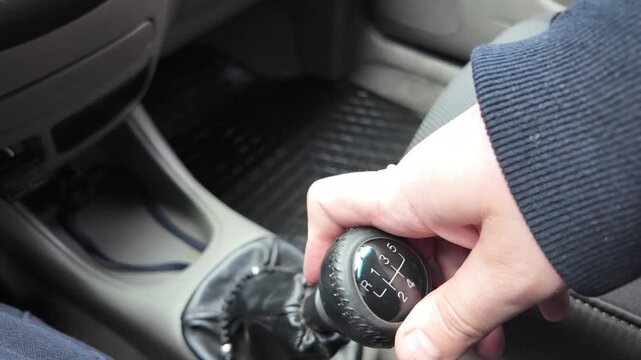 Close-up of a driver's hand shifting gears on a black leather manual transmission stick inside a car. A male hand holds the gearstick, controlling the vehicle. Automotive interior details.