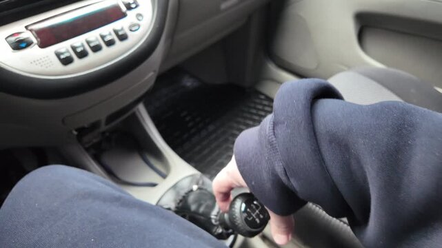 Close-up of a driver's hand shifting gears on a black leather manual transmission stick inside a car. A male hand holds the gearstick, controlling the vehicle. Automotive interior details.