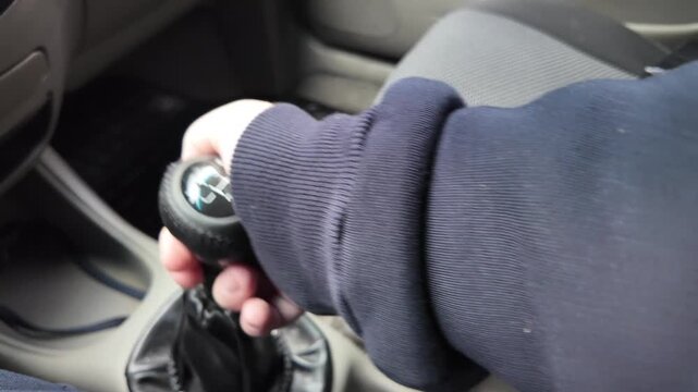 Close-up of a driver's hand shifting gears on a black leather manual transmission stick inside a car. A male hand holds the gearstick, controlling the vehicle. Automotive interior details.