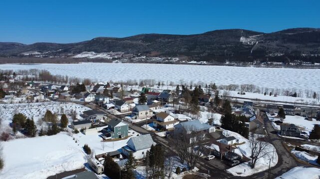 Aerial winter drone footage over Lac-au-Saumon shows snow‑covered homes, a frozen textured lake, and the Appalachian mountains under a clear blue sky in the Matapedia Valley, Quebec, filmed in 2026.