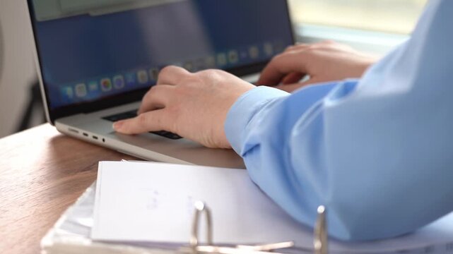 Moscow, March 20 2026, Close-up of hands typing on laptop keyboard. Light blue shirt sleeve visible. Wooden desk surface holds papers. Pens rest on documents. Natural light setting