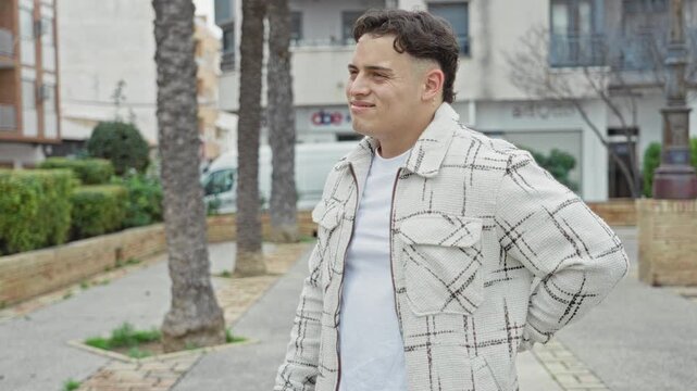 Young hispanic man smiling with hand behind back, wearing checked jacket and white tee on street near palm trees and planters; urban casual contentment.
