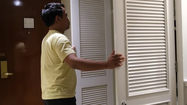 Southeast Asian man standing beside an open wardrobe with hands on head in a hotel room, showing stress, confusion, or frustration.