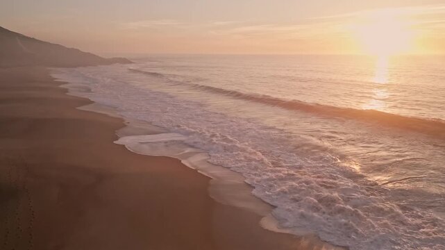 Stunning drone footage at Praia do Salgado near Nazare, Portugal. The video captures the golden hour sunset over the Atlantic coast, featuring breaking waves on a wide, empty sandy beach
