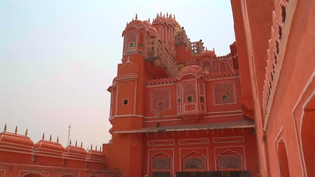 A static shot of the rear facade of the Hawa Mahal (Palace of Winds), featuring its distinctive pink sandstone walls, ornate domed pavilions, and traditional Rajput windows under a bright sky.