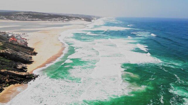 Filmed at Foz do Arelho in the Oeste region of Portugal, this drone shot captures the Atlantic ocean meeting the Obidos lagoon, featuring white sand, rugged cliffs, and crashing turquoise waves