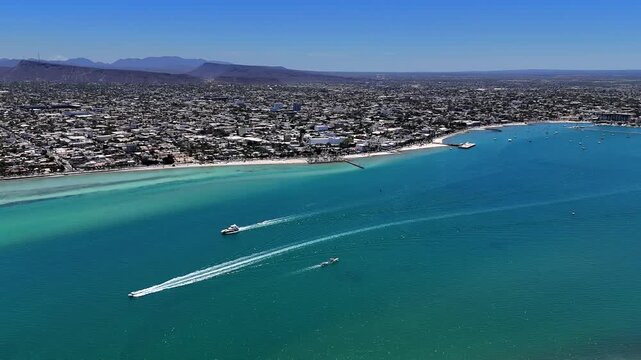 4K drone aerial over the ocean toward La Paz, showing the malec&oacute;n with a yacht and boats moving across bright blue water on a sunny day.