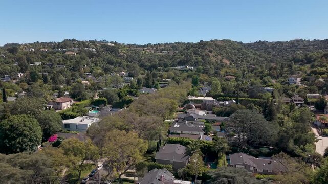 Aerial view of Sherman Oaks residential neighborhood with tree-lined streets, modern homes and rooftops nestled in green hills under clear blue sky in sunny Los Angeles, California. Wide shot.