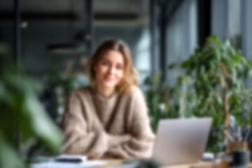 Fototapeta premium Blurred Young woman working on laptop in modern office, smiling, natural light, cozy sweater