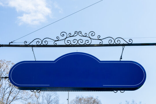 a blank blue hanging sign with a decorative wrought iron bracket, suspended against a clear light blue sky with faint white clouds, providing an empty mockup space for custom text or logos